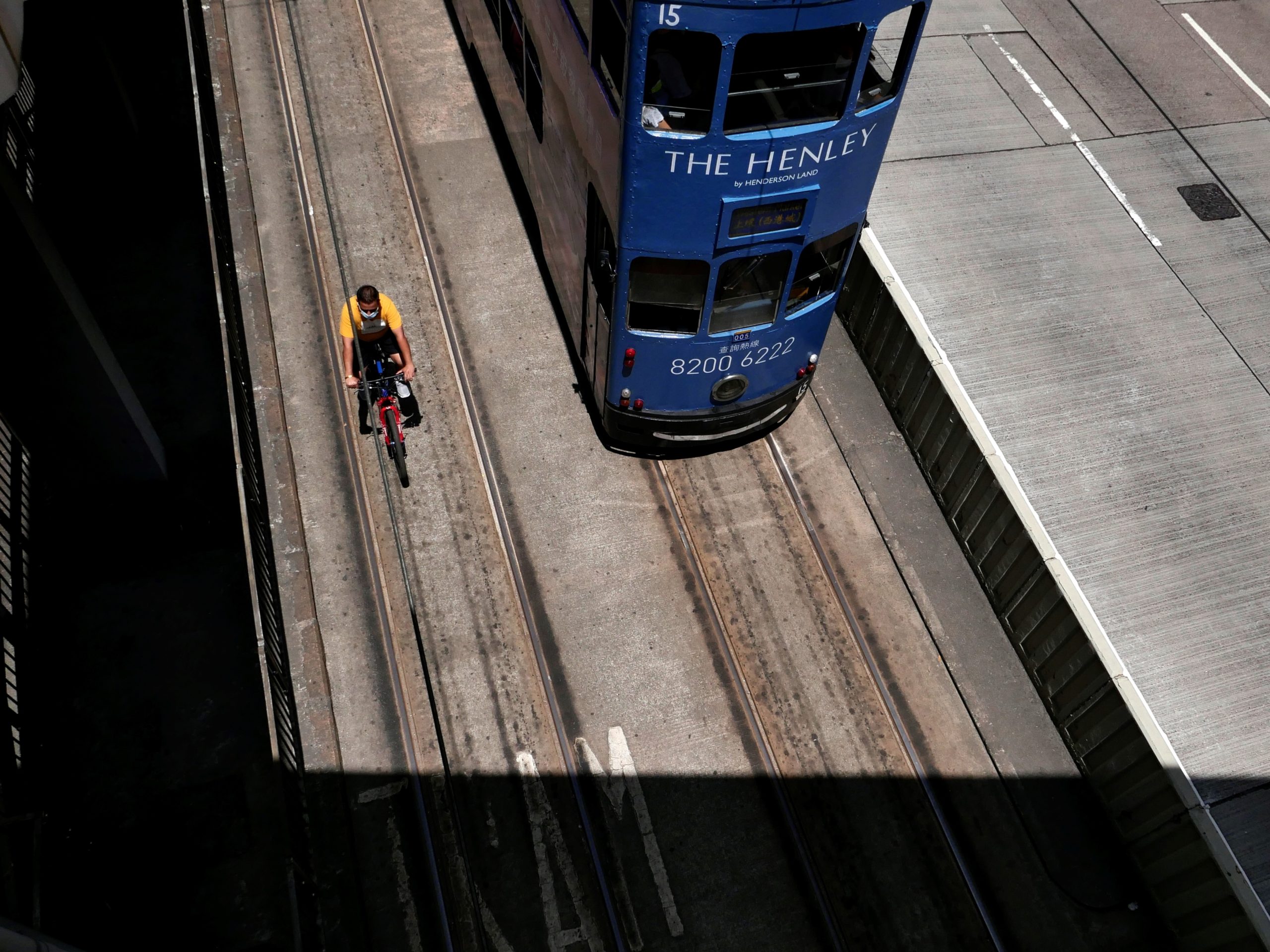 A blue-colored double decker tram driving by a cyclist in a yellow shirt, clearly blatant of all traffic regulations.